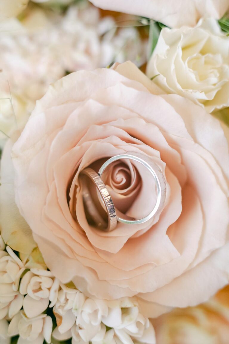 Close up of wedding rings elegantly placed atop a blush rose, surrounded by delicate floral arrangement.
