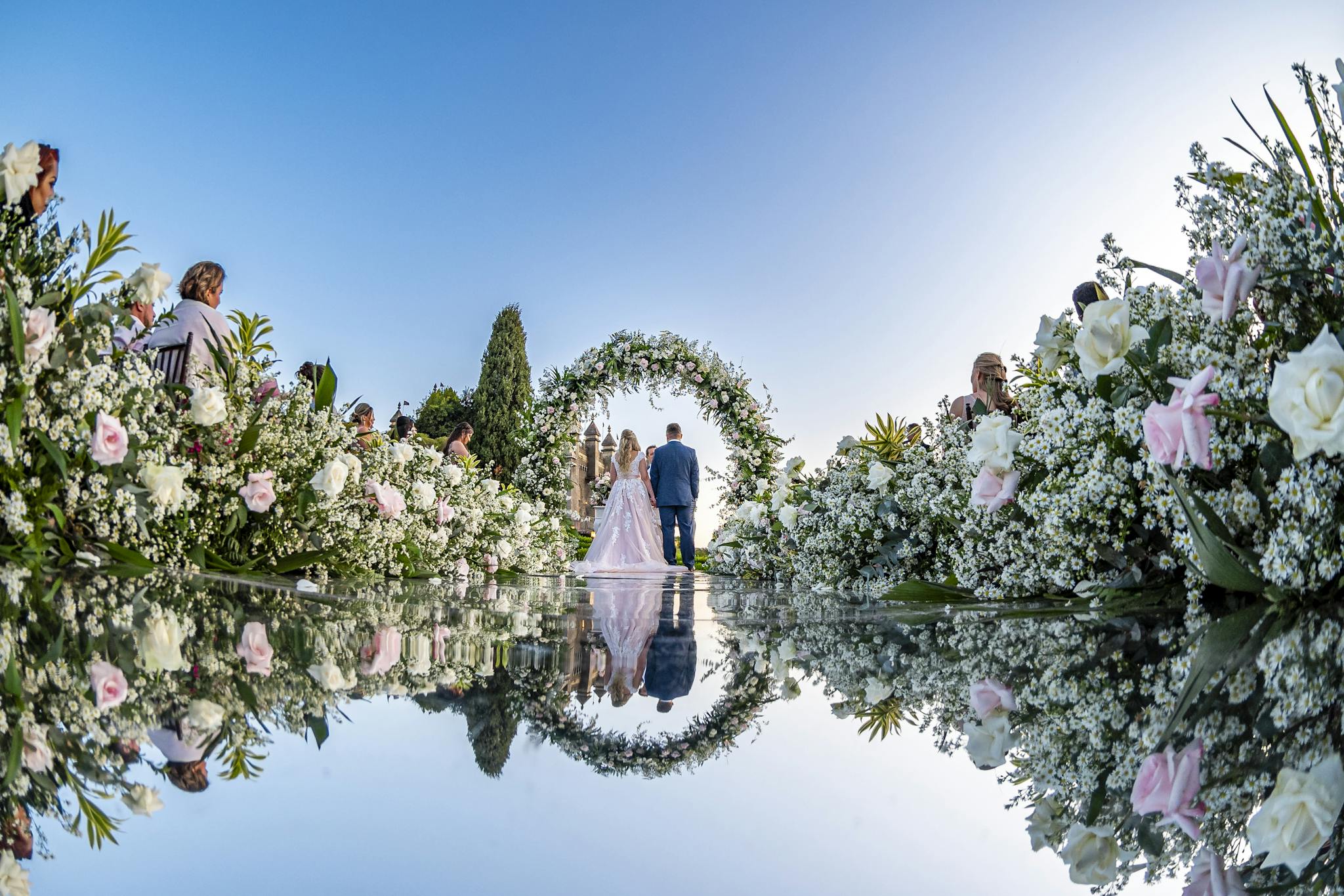 A romantic outdoor wedding ceremony featuring a floral arch, bride and groom reflection, and guests in a garden setting.